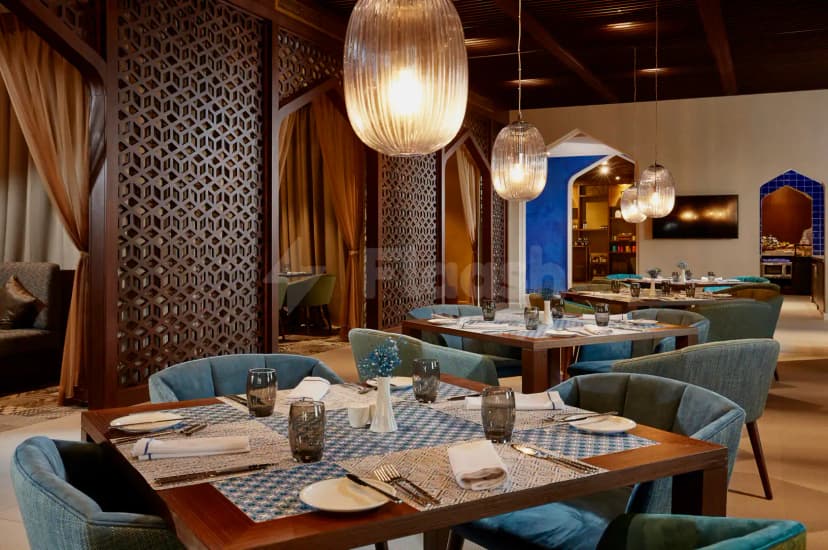 Restaurant at Shaza Riyadh featuring wooden tables with patterned runners, blue upholstered chairs, and hanging glass pendant lights