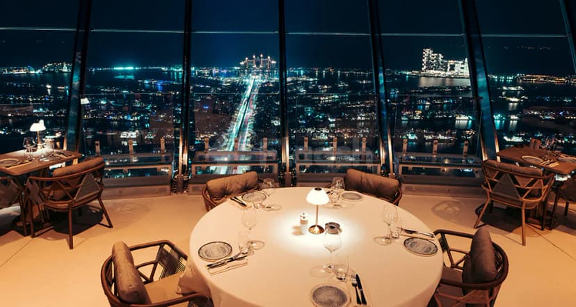 Restaurant at Coucou Dubai with a round set table and floor-to-ceiling windows overlooking the illuminated Dubai waterfront
