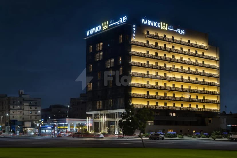 Hotel facade of Warwick Hotel Jeddah at night, featuring illuminated signage in English and Arabic, decorative balcony railings