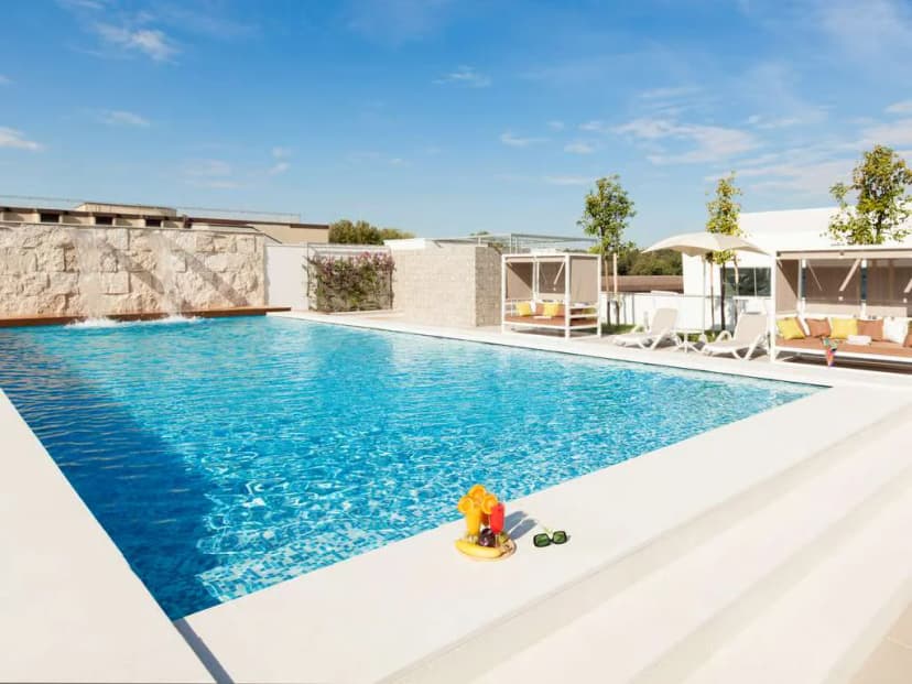 Rectangular swimming pool at Adagio Jumeirah Village Triangle in Dubai, featuring white tiled edges, a stone wall