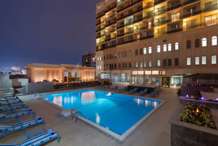 Rectangular illuminated swimming pool at Treffen House Doha with tiled deck, lounge chairs, and beige multi-story building facade