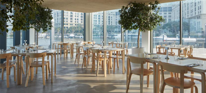 Restaurant at Jameel Arts Centre in Dubai with wooden tables, indoor trees, and large windows overlooking the waterfront