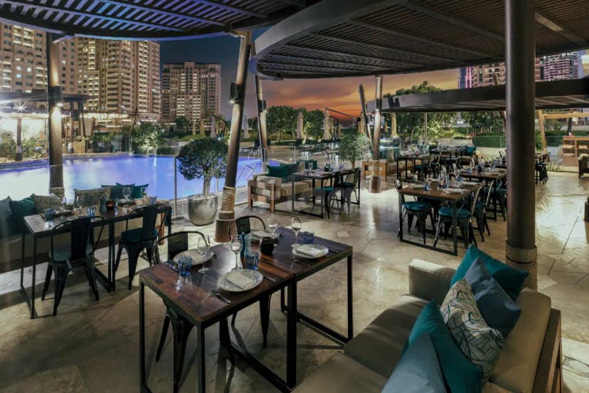 Rooftop terrace at The Kitchen Doha featuring wooden tables, black metal chairs, and a pool view with city