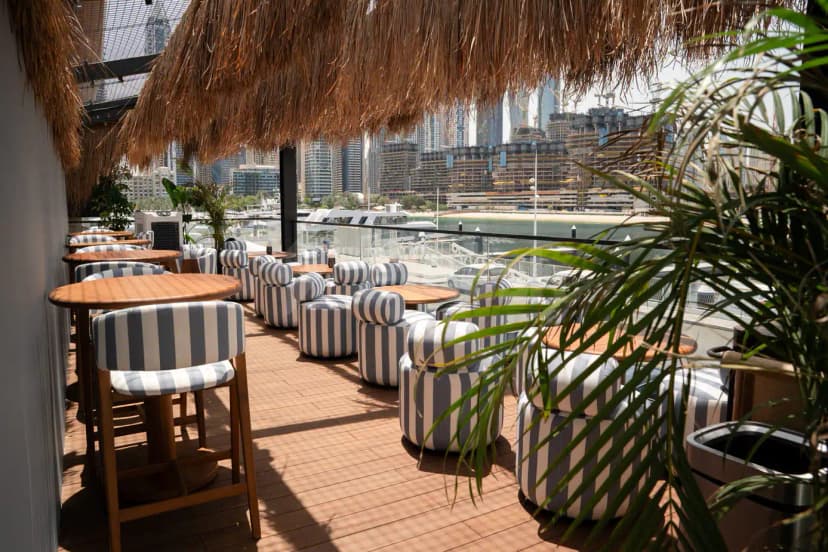 Rooftop terrace at Bar Du Port Dubai featuring a thatched roof, wooden deck, striped seating, and a waterfront