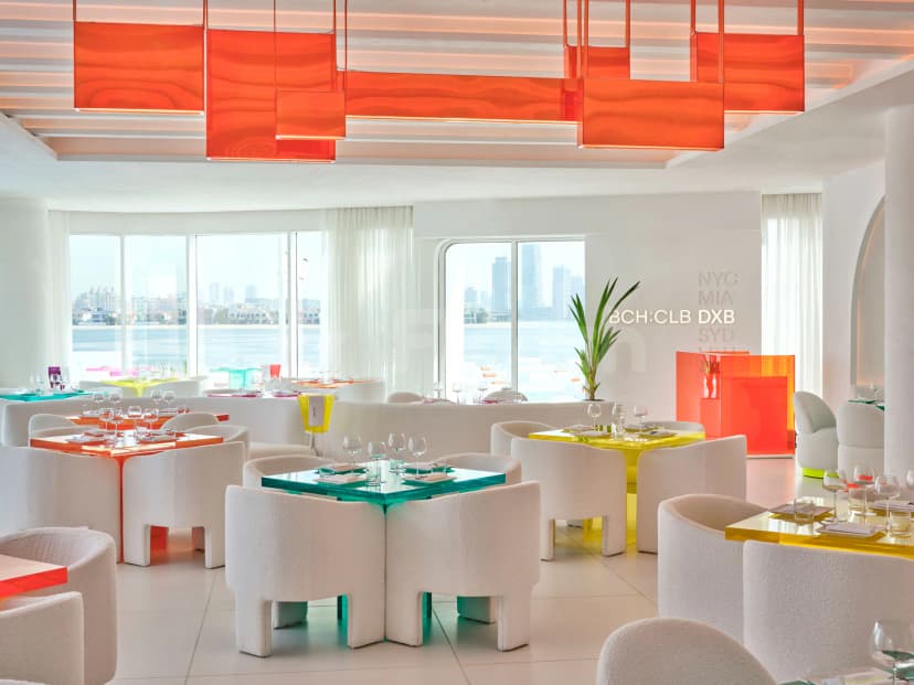 Restaurant interior at BCH CLB Dubai featuring white boucle chairs, colorful acrylic tables, and large waterfront windows