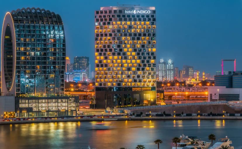 Hotel Indigo Dubai facade at dusk with illuminated windows and curved glass structure beside waterfront with yachts