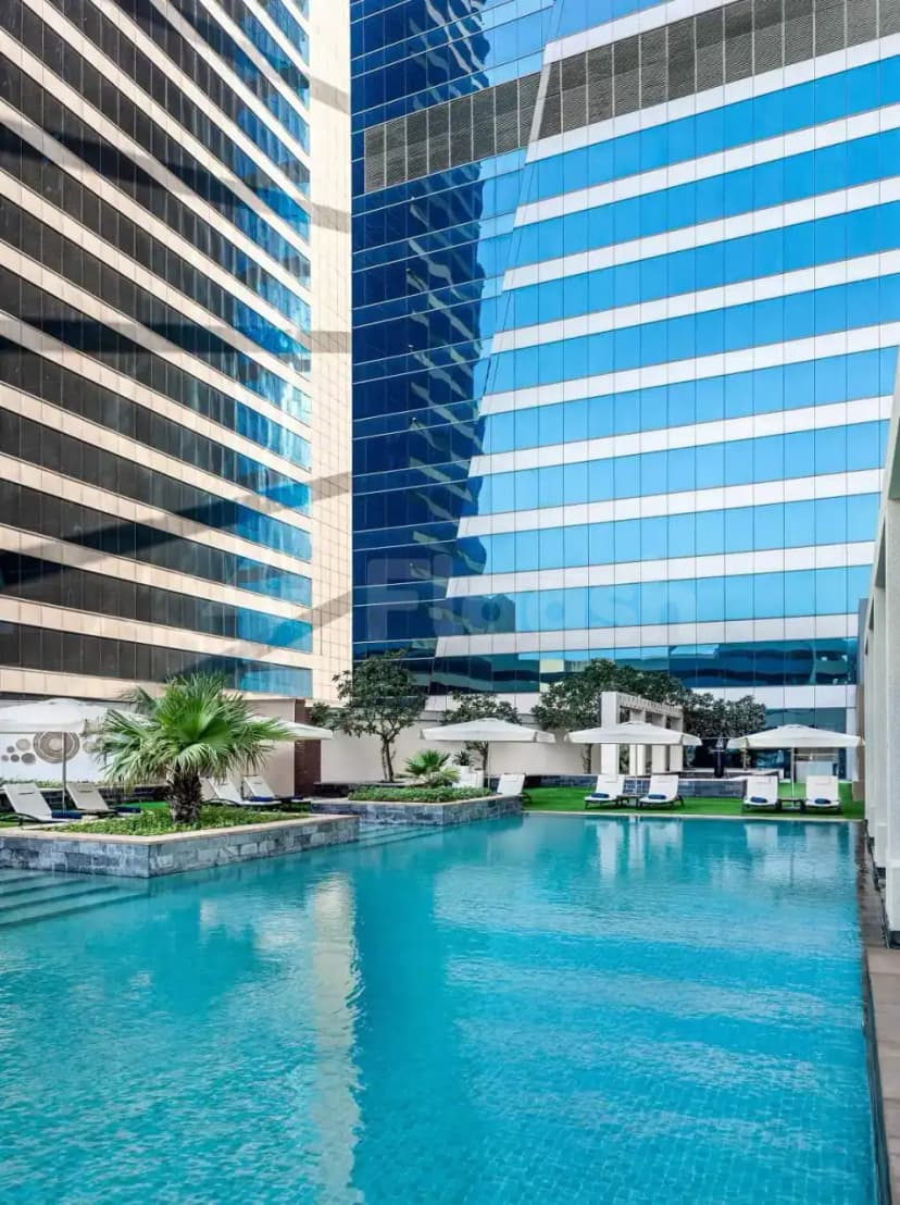 Rectangular swimming pool at The H Hotel Dubai with turquoise water, white lounge chairs, and glass-facade buildings nearby