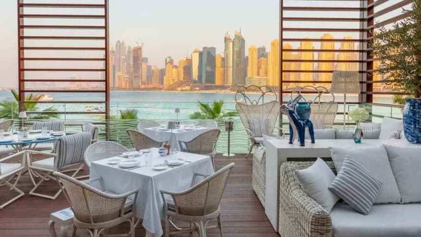 Rooftop terrace at Alici Restaurant Dubai featuring white tablecloths and wicker chairs with a city skyline and water