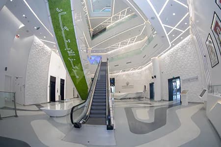 Entrance hall of Sustainability & Innovation Centre Dubai featuring a central escalator and a green Arabic-script banner against white