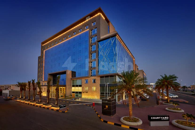 Hotel facade of Courtyard Jubail in Al Jubail featuring blue glass幕墙, geometric patterns, warm LED lighting, and palm