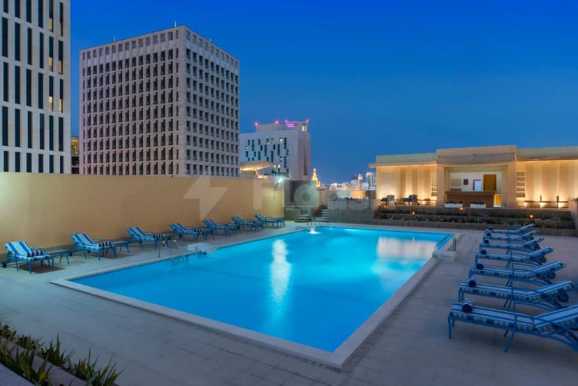 Rectangular swimming pool at Treffen House Doha with blue illuminated water, tiled deck, and lounge chairs under evening