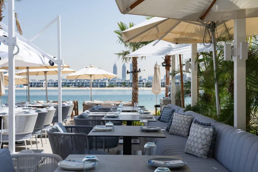 Outdoor dining area at Bebabel The Palm Restaurant in Dubai with white umbrellas, palm trees, and a waterfront