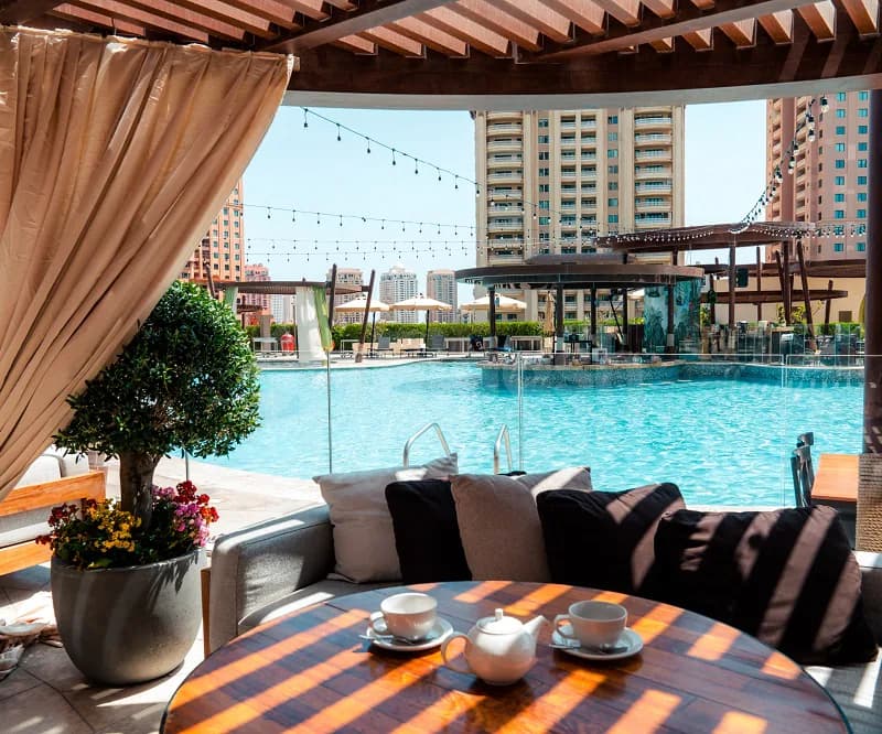 Rooftop terrace at The Kitchen Doha featuring a wooden pergola, beige curtain, and view of a blue pool
