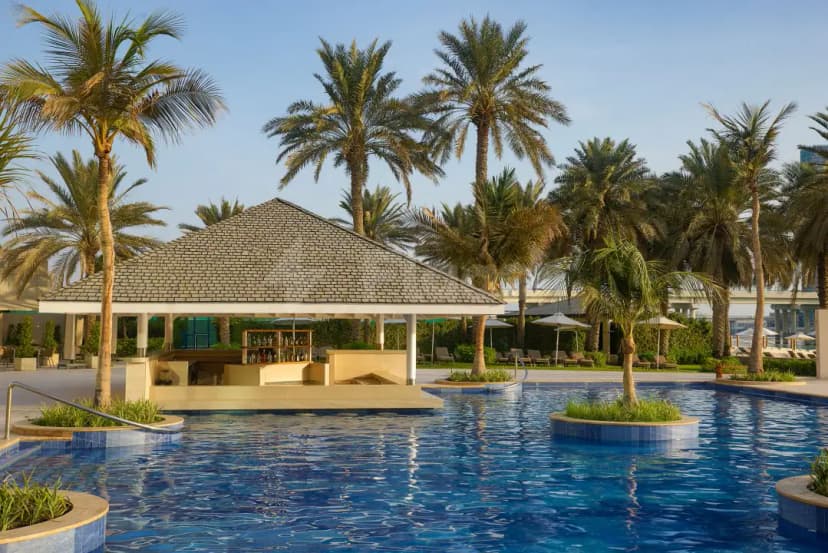 Swimming pool at Brauhaus Restaurant Abu Dhabi featuring a thatched-roof poolside bar and circular planters with palm trees