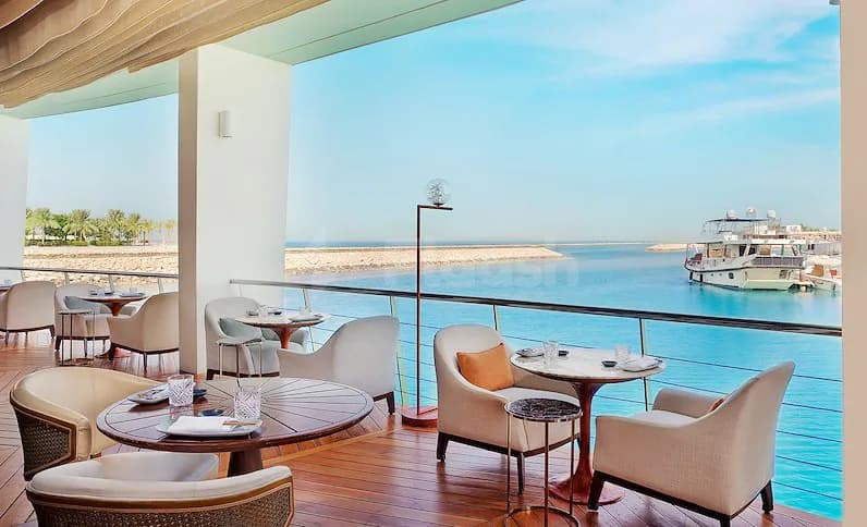 Rooftop terrace at B Lounge Doha featuring wooden deck flooring, beige armchairs, round tables, and a sea view