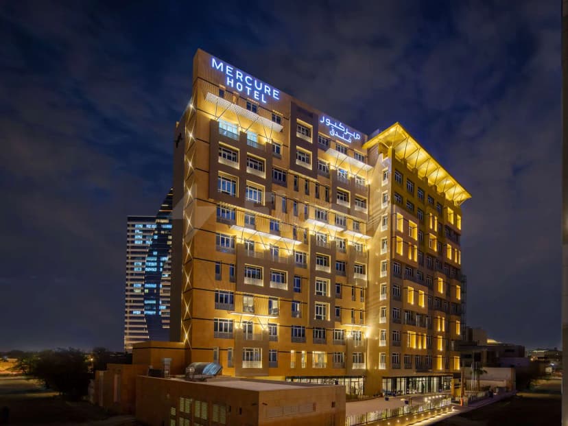 Hotel facade of Mercure Riyadh Al Anoud at night with illuminated blue signage and recessed lighting on beige