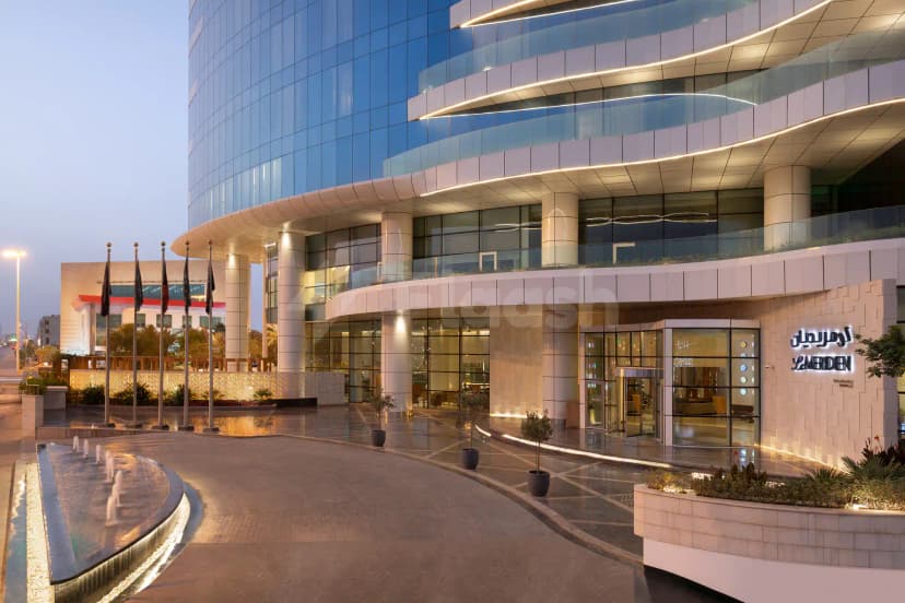 Hotel facade of Le Meridien Riyadh featuring a curved glass exterior, illuminated signage, and a water fountain