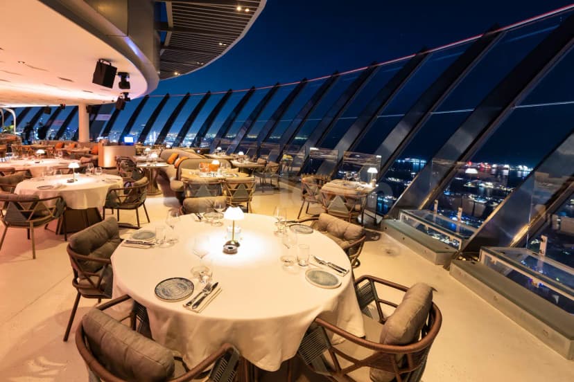 Rooftop restaurant Coucou Dubai with round dining tables, white tablecloths, and large angled glass windows overlooking the night
