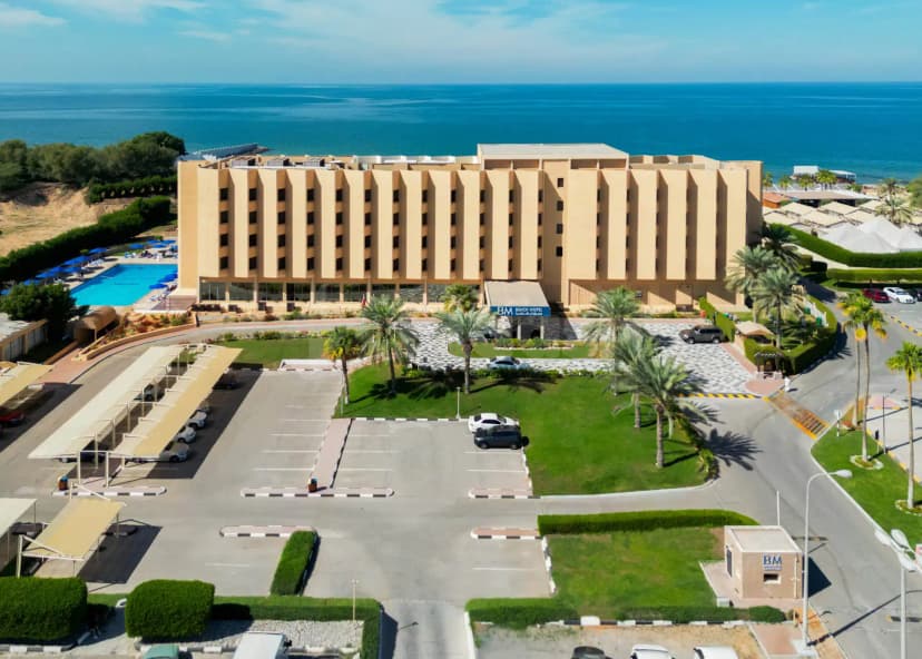 Hotel facade of BM Beach Resort Ras Al Khaimah with palm trees, green lawn, and ocean view