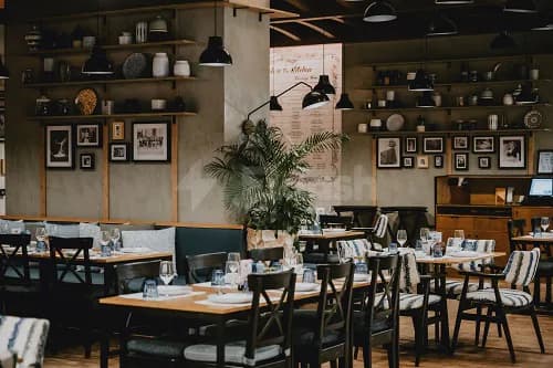 Restaurant interior at The Kitchen Doha featuring wooden tables, black chairs with striped upholstery, and a potted palm