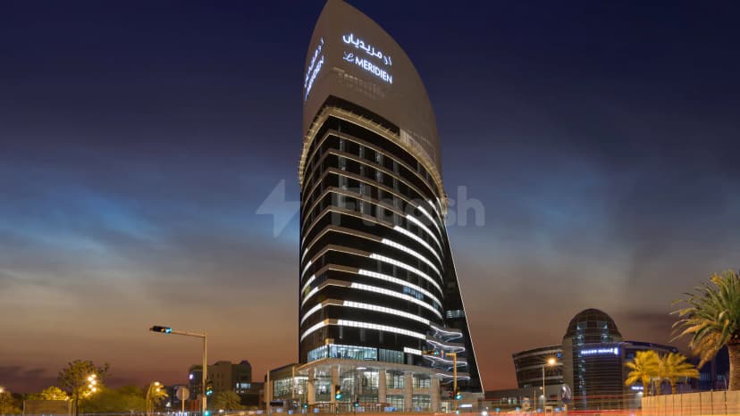 Hotel facade of Le Meridien Riyadh at dusk, featuring curved glass structure, illuminated Arabic and English signage