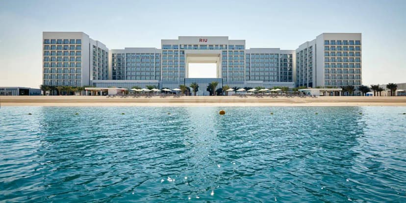 Hotel facade of Riu Dubai featuring a central archway, red RIU logo, palm trees, and a sandy beach