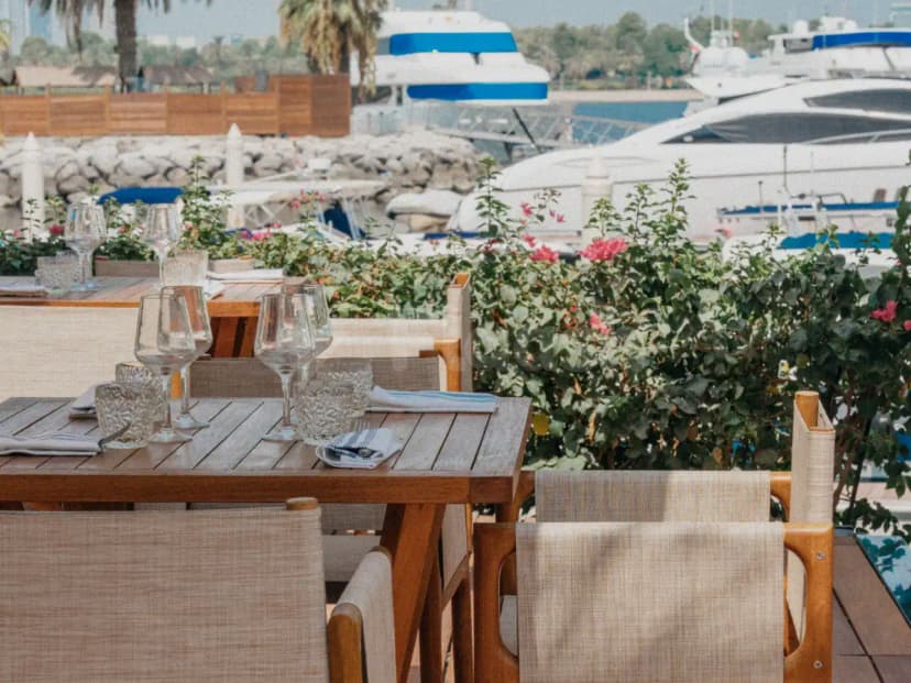 Terrace at Twiggy Dubai with wooden tables, beige fabric chairs, and potted bougainvillea overlooking white yachts