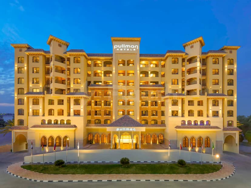 Hotel facade of Pullman Ras Al Khaimah with illuminated signage, arched windows, and a circular driveway featuring flagpoles