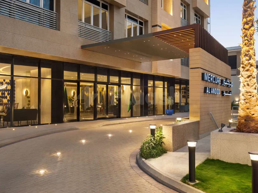 Hotel facade of Mercure Riyadh Al Anoud featuring glass entrance, stone cladding, illuminated sign, and a palm tree
