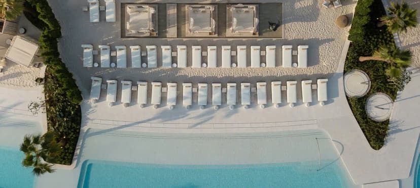 Swimming pool at San Beach Dubai with light blue water, white sun loungers on sand, and palm trees