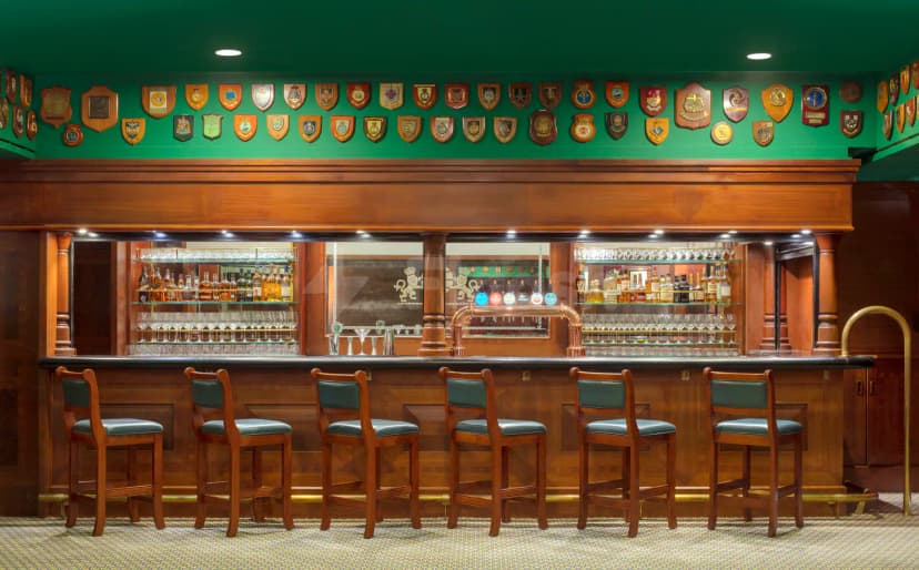 Bar counter at Theatre by QE2 in Dubai featuring wooden paneling, green leather stools, and shield plaques