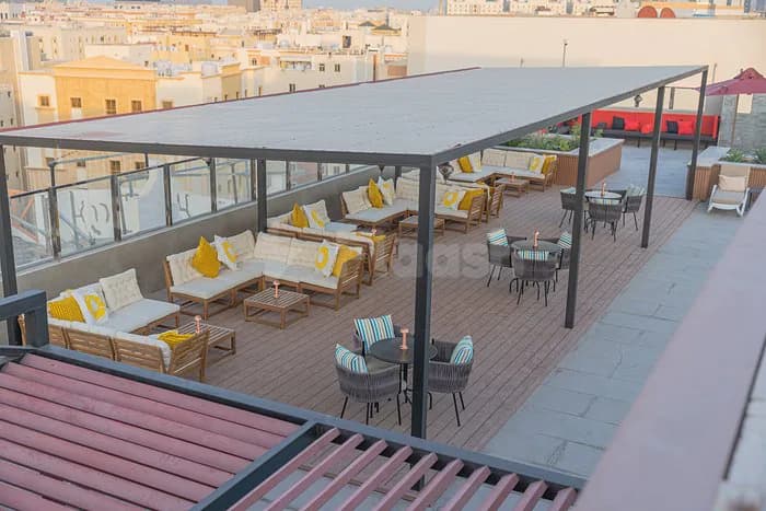 Rooftop terrace at Momento Lounge Doha featuring a metal pergola with white canopy, wooden deck, and yellow cushioned
