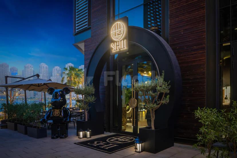 Arched entrance of Shi Restaurant Dubai Marina with illuminated sign, glass doors, potted olive trees, and black bear