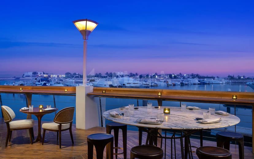 Rooftop terrace at B Lounge Doha featuring a marble-top table and wooden deck overlooking a marina with yachts
