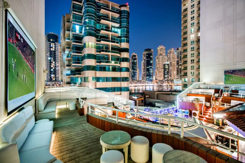 Rooftop terrace at Marina Byblos Hotel Dubai featuring wooden decking, white sofas, outdoor screens, and a city skyline