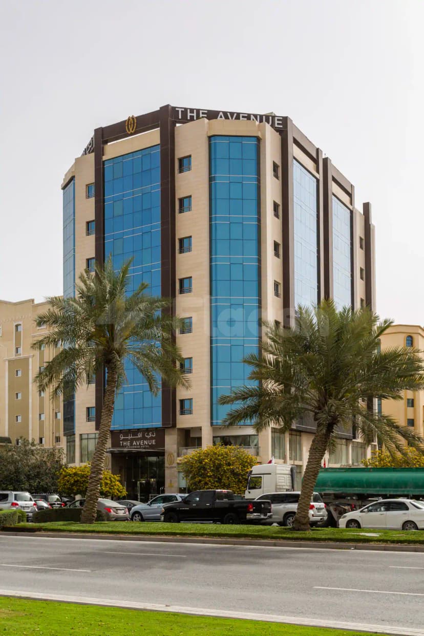 Hotel facade of The Avenue Hotel in Doha featuring blue glass panels, beige stone cladding, and palm trees
