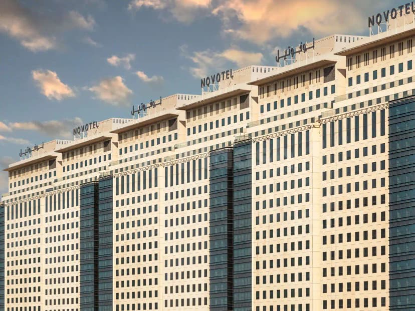 Hotel facade of Novotel Thakher in Makkah featuring beige concrete, dark glass vertical strips, and bilingual signage against