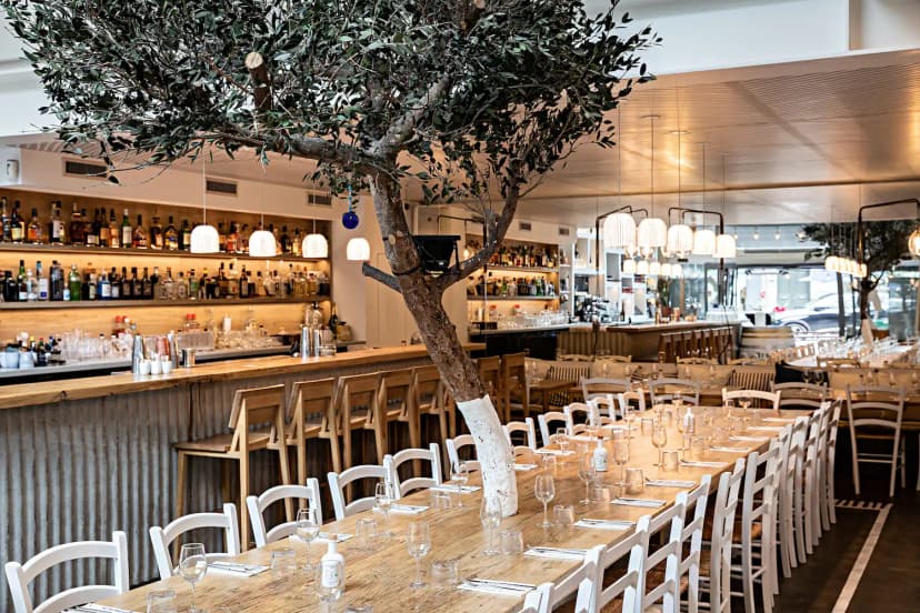 Restaurant événementiel chaleureux avec grandes tablées, bar central et oliviers intérieurs